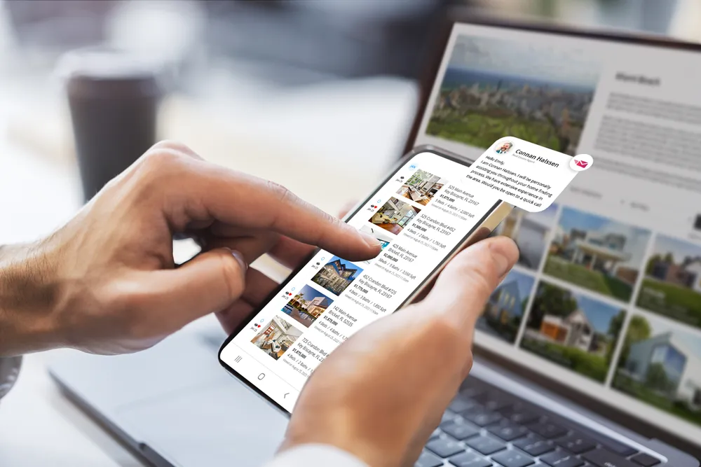 A person’s hands holding a smartphone that shows a list of home idx listings and a chat message from a real estate agent. In the background, a laptop screen also displays property information.