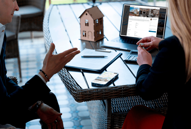 male real estate agent talking to a woman client sitting on chairs in front of a table, building a relationship and positioning himself as an expert on his niche area detailing property information using a laptop.